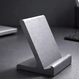 May include: A matte silver-coloured metal phone stand with an angled back support and a small front ledge to hold a mobile device in place. The stand features a smooth, solid construction with a clean, uniform finish and a stable base design.