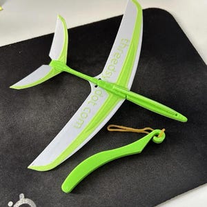 May include: A lime green and white toy glider airplane featuring the text "threedsy dot com" printed on the main wing. The set includes a separate lime green plastic launching handle with a small tan rubber band attached to the hook.