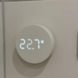 May include: A circular white digital thermostat mounted on a wall, featuring a glowing white LED display that shows the numerical value 22.7. The device has a smooth, matte finish and a simple, clean design with no visible buttons or markings.