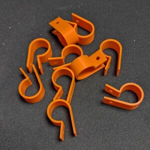 May include: A collection of orange plastic cable clamps. The clamps are U-shaped with a flat base and a small hole for mounting. They are scattered on a dark gray surface.
