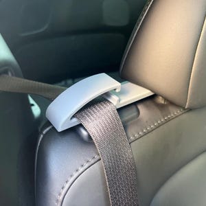 May include: A light gray car seat belt adjuster clip installed on a black car seat. The seat belt is black and is threaded through the clip. The car seat is made of black leather.