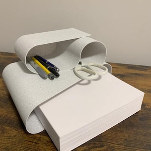 Peut inclure: Un organisateur de bureau gris clair incurvé, fabriqué dans un matériau flexible et moucheté. Il comporte deux compartiments ouverts contenant trois stylos et une paire de ciseaux blancs, avec une pile de papier blanc dans la section inférieure.