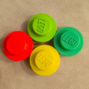 Puede incluir: Cuatro bloques de construcción LEGO circulares de plástico en rojo brillante, verde lima, verde bosque y amarillo. Cada pieza presenta un único perno elevado en la superficie superior con el logotipo LEGO en relieve visible en el centro de cada perno.