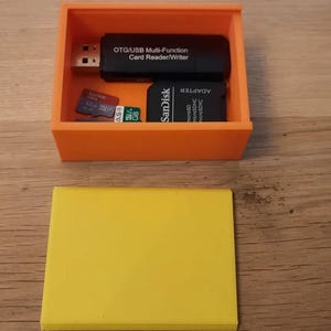 May include: An orange rectangular storage box containing a black OTG/USB multi-function card reader/writer, a black SanDisk adapter, and two memory cards. A yellow rectangular object is below the box. The items are on a wooden surface.