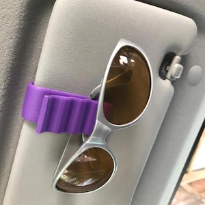 May include: A purple, wavy-textured plastic clip attached to a light gray car sun visor, holding a pair of silver-framed sunglasses with brown tinted lenses. The clip features a ridged design to securely grip the eyewear frame in place.