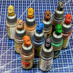May include: A collection of miniature paint bottles with colorful, sculpted monster head caps. The bottles are arranged on a blue grid surface. The caps are various colors, including yellow, orange, blue, green, red, and purple.