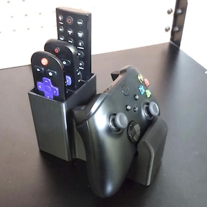 May include: A black plastic storage stand featuring a vertical compartment holding two remote controls, one with a purple directional pad, and a front-facing cradle supporting a black wireless game controller with colored action buttons.
