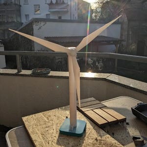 以下が含まれることがあります： 円筒形のタワーの頂上にある中央ハブに3枚の長く細いブレードが取り付けられた、白い風力タービンの模型。タワーは正方形のティールカラーのベースに取り付けられています。構造は滑らかでマットな質感で仕上げられています。