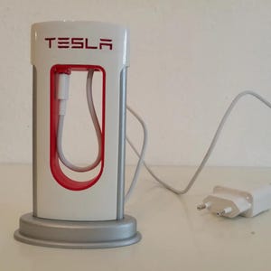 May include: A white and silver Tesla desktop charging station featuring a red interior accent and the Tesla logo printed in red at the top. The device includes a white charging cable and a white wall plug adapter with two metal prongs.