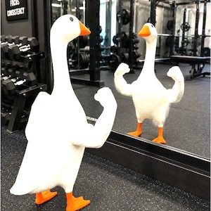 May include: A white goose figurine with orange feet and an orange beak, featuring muscular human-like arms flexed in a bodybuilding pose. The goose stands before a mirror, reflecting its muscular arms and posture in the gym setting.