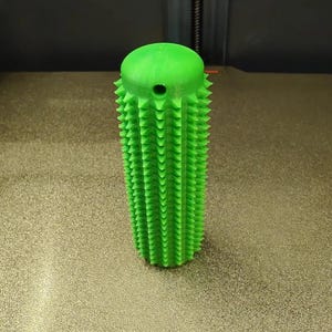 May include: A bright green cylindrical object with a textured surface covered in small, uniform spikes. The top of the cylinder is rounded and features a single, small, circular hole positioned near the upper edge of the spiked body.