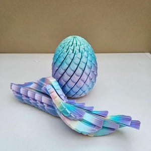 May include: A 3D-printed dragon egg and tail in shades of blue, purple, and white. The egg is oval-shaped with a textured surface. The tail is segmented and curved, with a rainbow gradient.