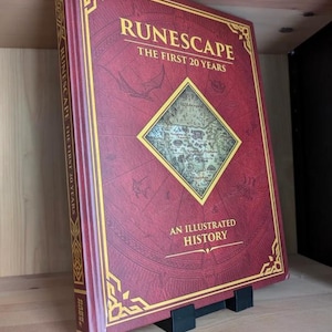 May include: A hardcover book titled RuneScape: The First 20 Years, An Illustrated History. The cover is deep red with gold foil accents, featuring a central diamond-shaped map illustration and intricate geometric border designs on the front.