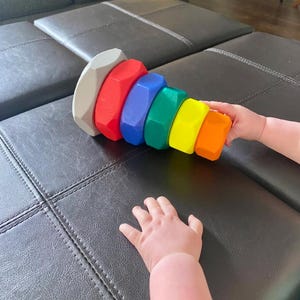 May include: A colorful wooden stacking toy with a gray top and a series of red, blue, green, yellow, and orange blocks. The toy is held by a child's hands. The toy is on a black leather surface.