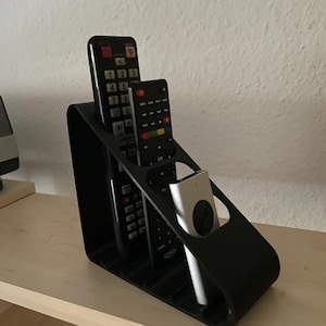 May include: A black plastic remote control organiser with a slanted design featuring three distinct slots. Two black rectangular remote controls are placed in the back slots, and a smaller silver remote control is positioned in the front slot.