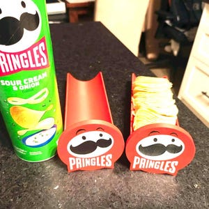May include: A green Pringles Sour Cream & Onion potato chip can next to two red plastic Pringles-branded chip holders. One holder is empty and the other is filled with a stack of light yellow potato chips, each featuring the Pringles logo.