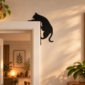 Può includere: Una decorazione da parete in metallo nero a forma di sagoma di gatto, progettata per essere montata sull'angolo di uno stipite di una porta. Il gatto è raffigurato in una posa di arrampicata, con le zampe anteriori sul bordo superiore e il corpo lungo il lato.