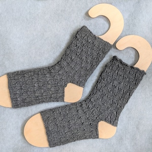 Puede incluir: Dos pares de calcetines de punto grises con un patrón de trenza, expuestos en perchas de calcetines de madera.