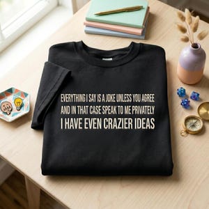 May include: A black crewneck sweatshirt featuring the white text: EVERYTHING I SAY IS A JOKE UNLESS YOU AGREE AND IN THAT CASE SPEAK TO ME PRIVATELY I HAVE EVEN CRAZIER IDEAS. The garment is displayed on a light wooden surface with various desk items.