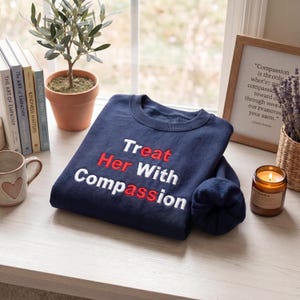 May include: A navy blue sweatshirt with the embroidered text "Treat Her With Compassion" in white and red. The sweatshirt is folded on a light-colored surface, with books, a candle, and a small potted plant in the background.