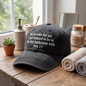 May include: A black baseball cap with the text "oh no coke for me I just wanted to be in this tiny bathroom with you ;-) " in white. The cap is sitting on a wooden surface.