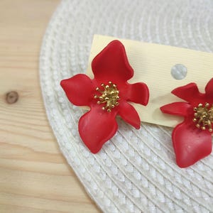Può includere: Un paio di orecchini a lobo a forma di fiore rosso brillante a cinque petali con finitura opaca. Il centro di ogni fiore è decorato con un gruppo di piccoli stami metallici color oro. Gli orecchini sono fissati su un cartoncino rettangolare.