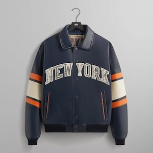 Puede incluir: Chaqueta universitaria azul marino con el texto "NEW YORK" en el pecho. La chaqueta tiene rayas crema y naranja en las mangas y un dobladillo acanalado negro. La chaqueta est&aacute; colgada de una percha negra.