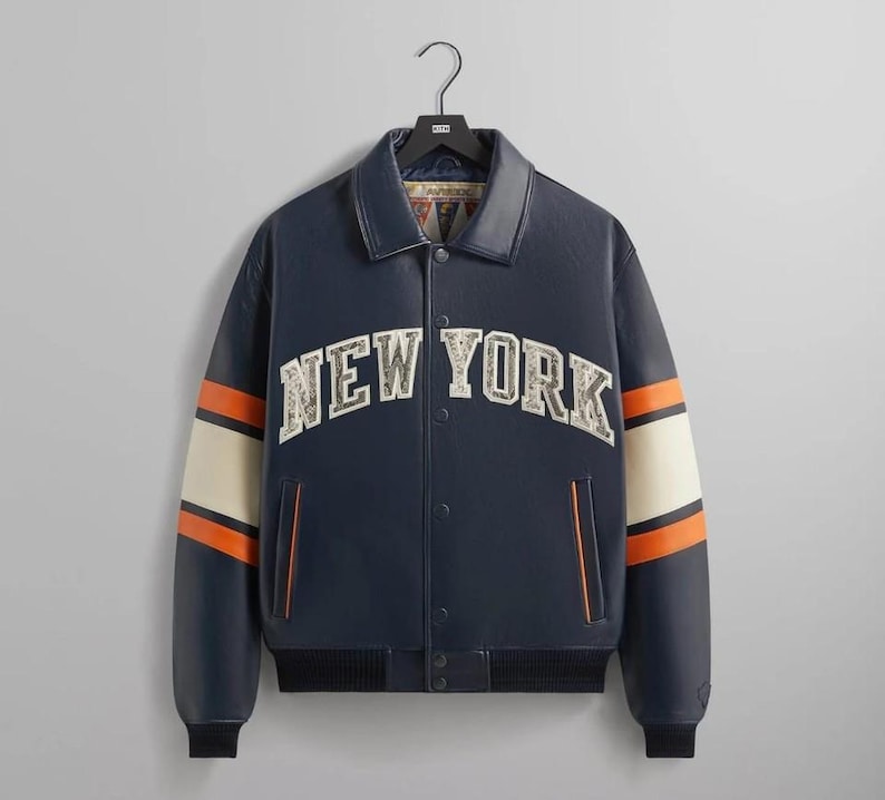 Puede incluir: Chaqueta universitaria azul marino con el texto "NEW YORK" en el pecho. La chaqueta tiene rayas crema y naranja en las mangas y un dobladillo acanalado negro. La chaqueta est&aacute; colgada de una percha negra.