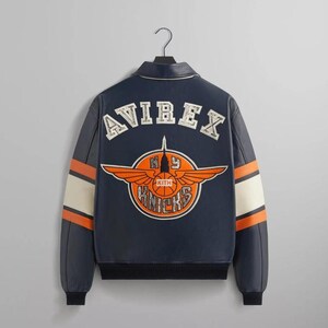 Puede incluir: Chaqueta de cuero azul marino con la palabra "AVIREX" en los hombros. La parte trasera presenta un gran logotipo naranja y blanco con las palabras "NY Knicks" y "KITH". Las mangas tienen rayas naranjas y blancas.