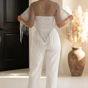 Peut inclure: Un châle argenté à perles et à franges est porté sur un haut blanc et un pantalon blanc. Le châle est drapé sur les épaules et a une forme triangulaire à l'avant. La tenue est complétée par des talons blancs.
