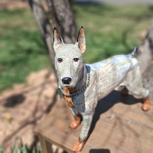 Peut inclure: Sculpture artisanale d'un chien au design unique. Le chien est recouvert d'imprimés de journaux, avec des accents marron et noirs sur les oreilles et les pattes. Il porte un collier en chaîne argentée. La sculpture est sur une surface en bois.