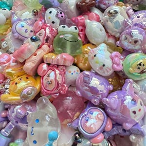 May include: A collection of assorted small plastic charms and beads in various pastel colours. The items feature various cartoon-inspired animal shapes, including frogs, cats, and bears, with glossy, iridescent, and translucent finishes.