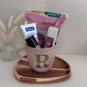 May include: A light pink ceramic mug with a gold letter R and the name Raquel printed on the front. Inside the mug are a tube of Elina hand cream, a Garnier sheet mask, a pink patterned nail file, and a tube of lip gloss with a black cap.