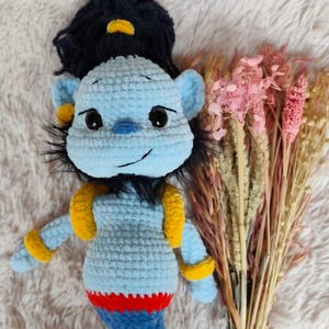 May include: A handmade blue crochet doll with black hair and yellow accents. The doll has a smiling face, large black eyes, and a red and blue bottom. The doll is lying on a white, fluffy surface next to a bouquet of dried flowers.