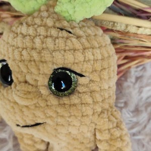 May include: A handmade, plush, light brown creature with large, glittery green and black eyes and a smiling mouth. The creature has a textured, fuzzy appearance and green leaves on top of its head. The background is blurred.