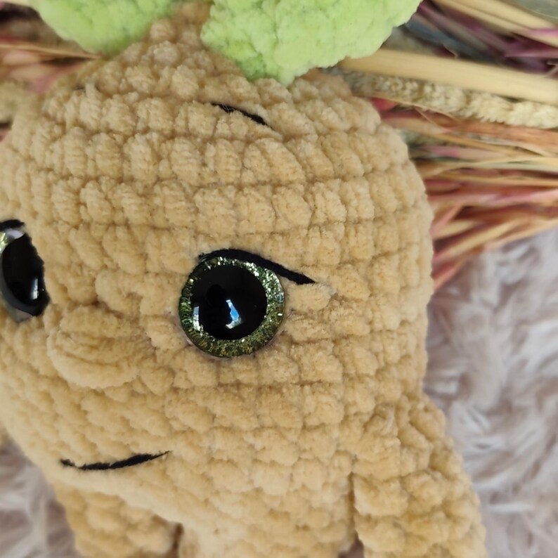 May include: A handmade, plush, light brown creature with large, glittery green and black eyes and a smiling mouth. The creature has a textured, fuzzy appearance and green leaves on top of its head. The background is blurred.