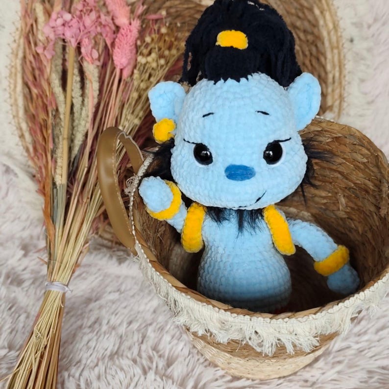 May include: A light blue plush genie doll with black hair and yellow accents sits in a woven basket. The doll has large black eyes and is made of soft, textured material. The basket has a brown handle and is decorated with white fringe.