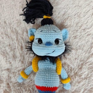 May include: A handmade crochet doll in the likeness of a genie. The doll is primarily light blue with yellow accents on the arms and ears. It has black hair and a red, blue, and yellow striped bottom.