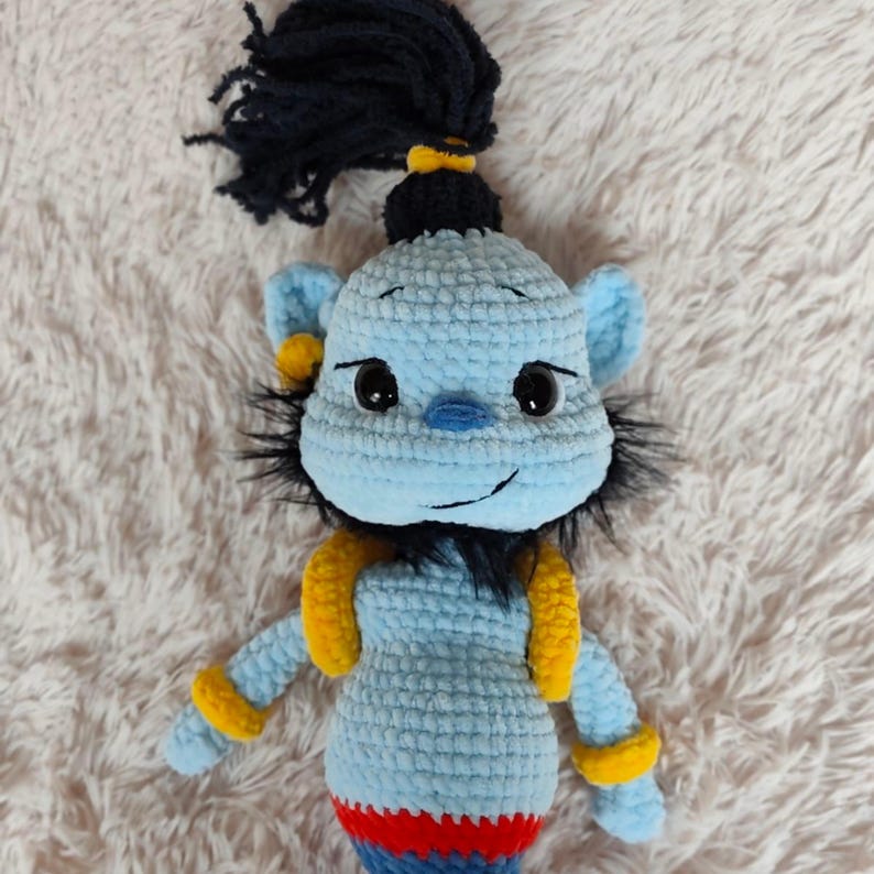 May include: A handmade crochet doll in the likeness of a genie. The doll is primarily light blue with yellow accents on the arms and ears. It has black hair and a red, blue, and yellow striped bottom.