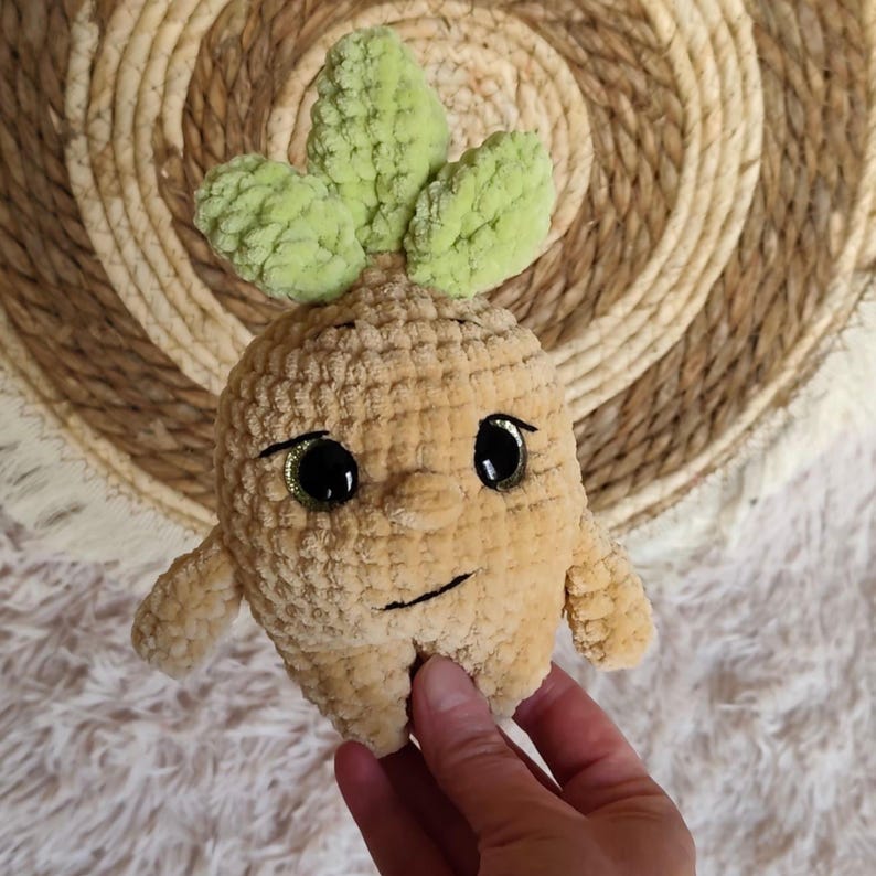 May include: A tan crocheted plush toy shaped like a root vegetable with a smiling face, large black eyes, and green leaves on top. The toy has small arms and is held by a hand.