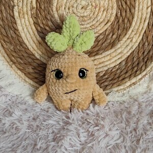 May include: A plush, tan-colored crochet doll resembling a mandrake plant, with green leaves, black eyes, and a stitched mouth. The doll has small arms and legs. The background is a woven, circular mat and a fluffy, white surface.