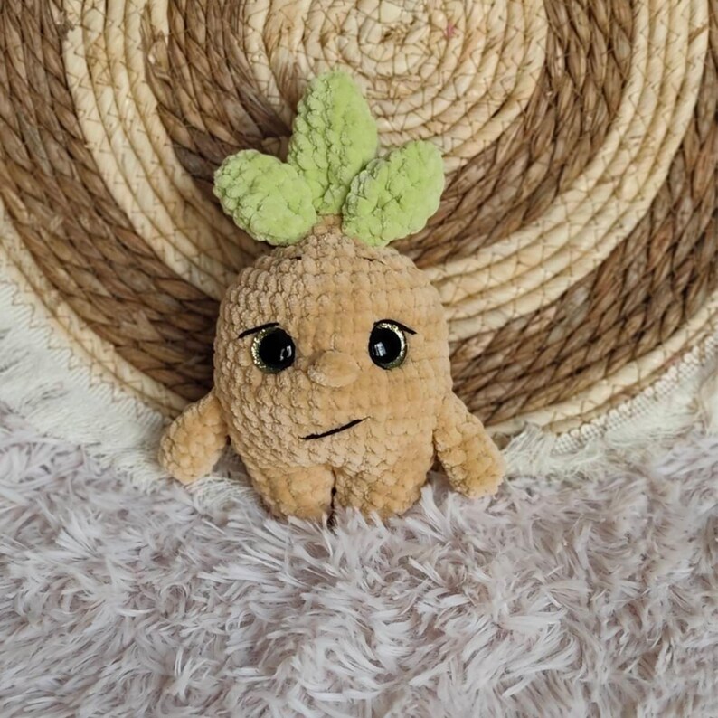 May include: A plush, tan-colored crochet doll resembling a mandrake plant, with green leaves, black eyes, and a stitched mouth. The doll has small arms and legs. The background is a woven, circular mat and a fluffy, white surface.