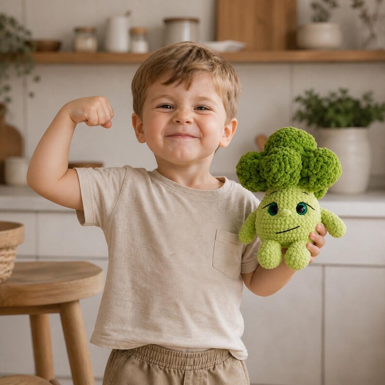 K&ouml;nnte beinhalten: Ein Kind l&auml;chelt und zeigt seinen Arm, w&auml;hrend es ein gr&uuml;nes, geh&auml;keltes Brokkoli-Spielzeug h&auml;lt. Das Spielzeug hat ein l&auml;chelndes Gesicht und kleine Arme und Beine. Das Kind tr&auml;gt ein beiges T-Shirt.