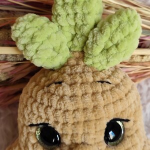 May include: A close-up of a crocheted plush toy with a light brown body and green leaves. The toy has large, shiny black eyes and a stitched mouth. The plush is made of soft, textured yarn.