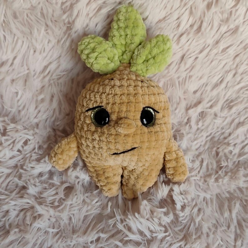 May include: A handmade plush toy in the shape of a root vegetable, featuring a tan body, green leaves, and large black eyes. The toy has a smiling face and small arms and legs, perfect for a child's toy collection.