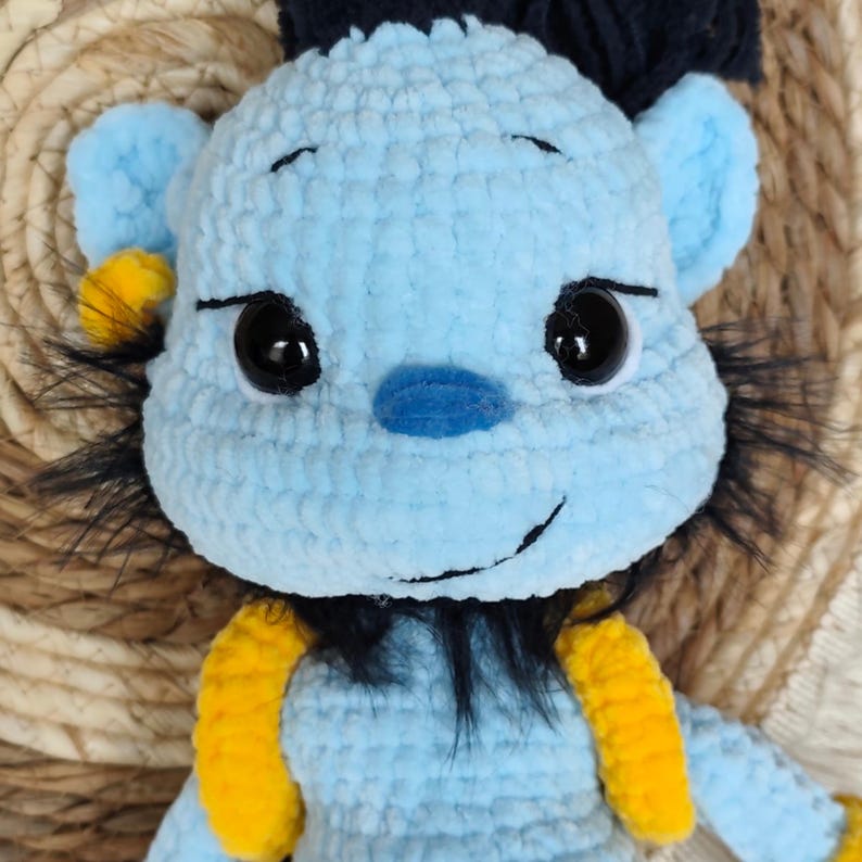 May include: A handmade, light blue crochet doll with black and yellow accents. The doll has large black eyes, a blue nose, and a smiling mouth. It features black hair and yellow straps. The doll is a soft, plush toy.