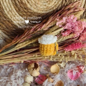 Może przedstawiać: Mały, szydełkowy kufel piwa z białą pianą i żółtym korpusem, o wysokości 5 cm. Tekst "crochet pattern" znajduje się nad kuflem. Tło obejmuje suszone kwiaty i elementy dekoracyjne.