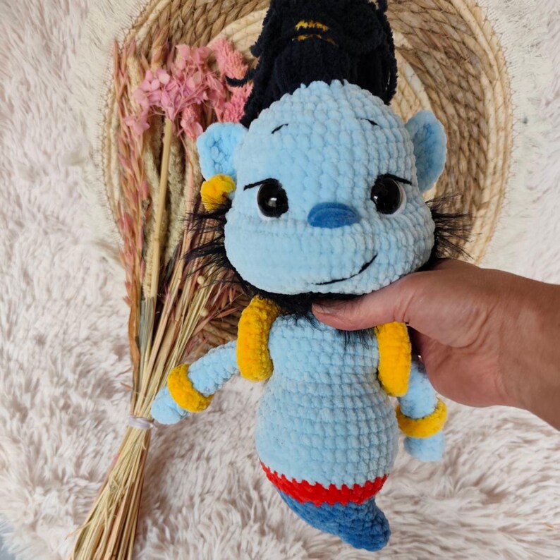 May include: A handmade blue crochet genie doll with black hair, yellow armbands, and a red and blue bottom. The doll has a smiling face with black eyes and a blue nose. The genie is a soft, plush toy.