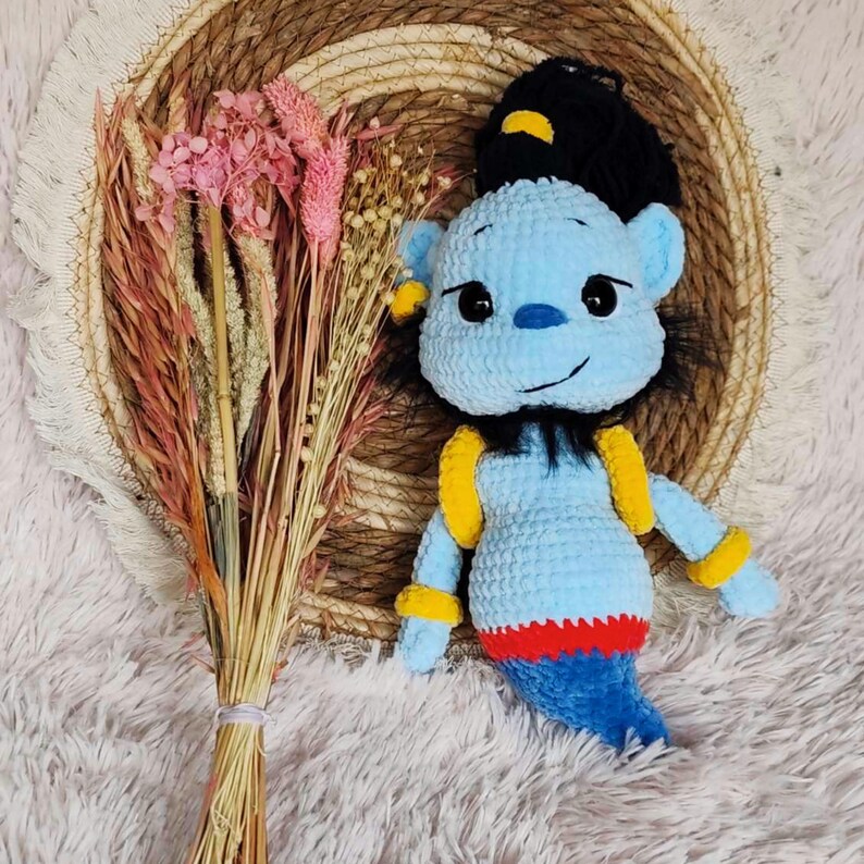 May include: A handmade blue crochet genie doll with black hair, yellow accents, and a red and blue tail. The doll is placed on a white fluffy surface next to a bouquet of dried pink and beige flowers.