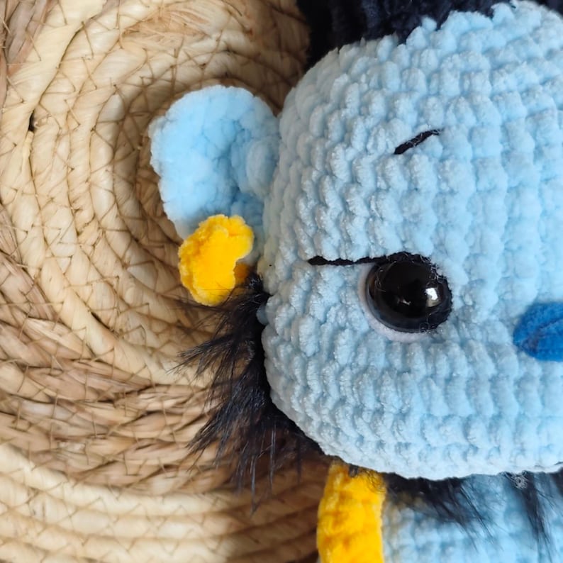 May include: A close-up of a light blue crocheted plush toy with black and yellow accents. The toy has a large black eye, a small blue nose, and a yellow ear. The background is a woven straw material.